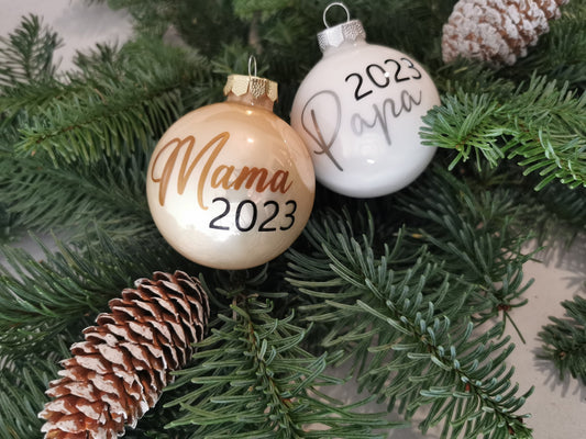 Weihnachtskugeln Mama 2023 und Papa 2023, personalisiert, aus glänzendem Glas in Gold und Weiß, dekoriert am Christbaum mit Tannenzweigen und Zapfen.