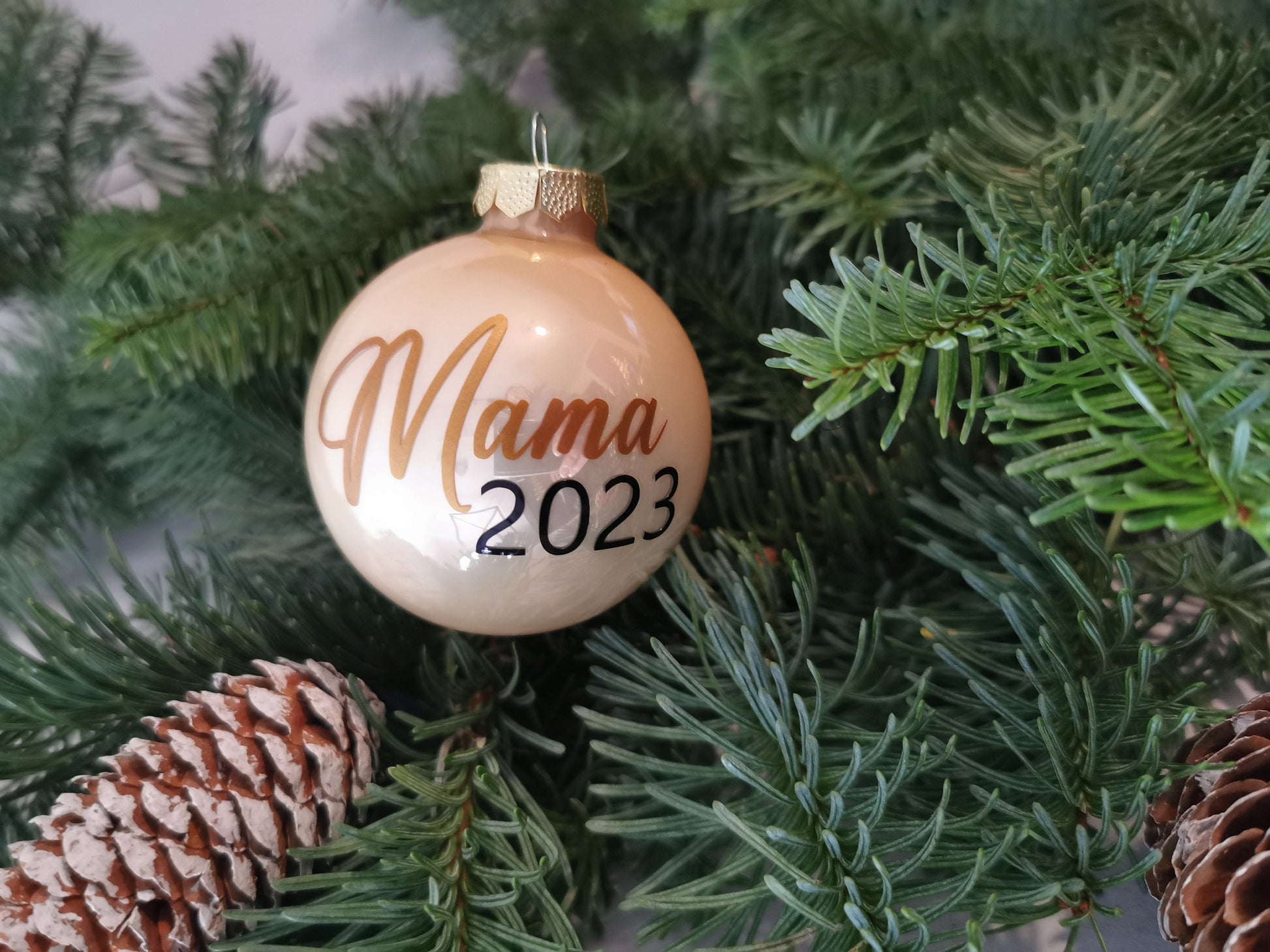 Weihnachtskugeln Mama 2023 und Papa 2023, personalisiert, aus glänzendem Glas in Gold und Weiß, dekoriert am Christbaum mit Tannenzweigen und Zapfen.