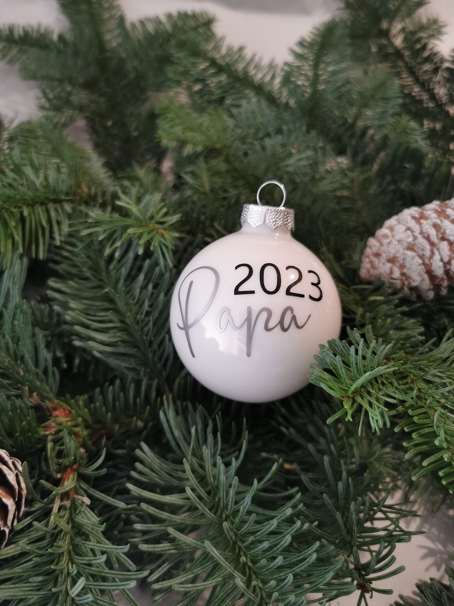 Weihnachtskugeln Mama 2023 und Papa 2023, personalisiert, aus glänzendem Glas in Gold und Weiß, dekoriert am Christbaum mit Tannenzweigen und Zapfen.