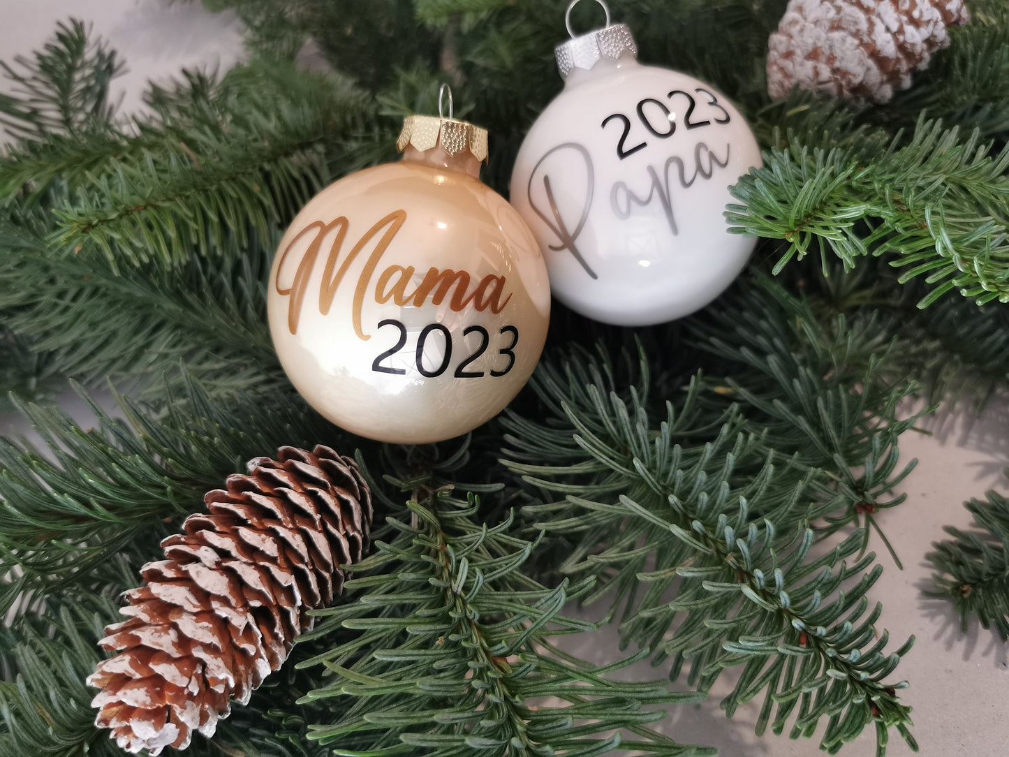 Weihnachtskugeln Mama 2023 und Papa 2023, personalisiert, aus glänzendem Glas in Gold und Weiß, dekoriert am Christbaum mit Tannenzweigen und Zapfen.