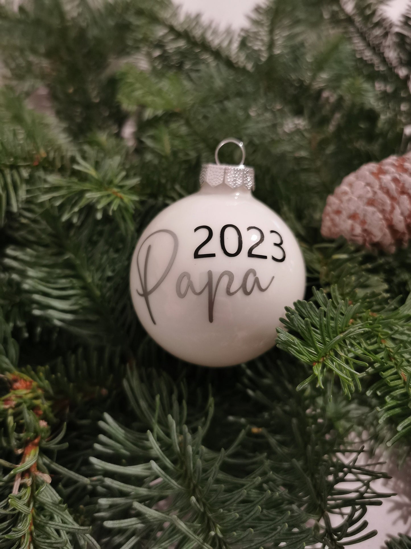 Weihnachtskugeln Mama 2023 und Papa 2023, personalisiert, aus glänzendem Glas in Gold und Weiß, dekoriert am Christbaum mit Tannenzweigen und Zapfen.