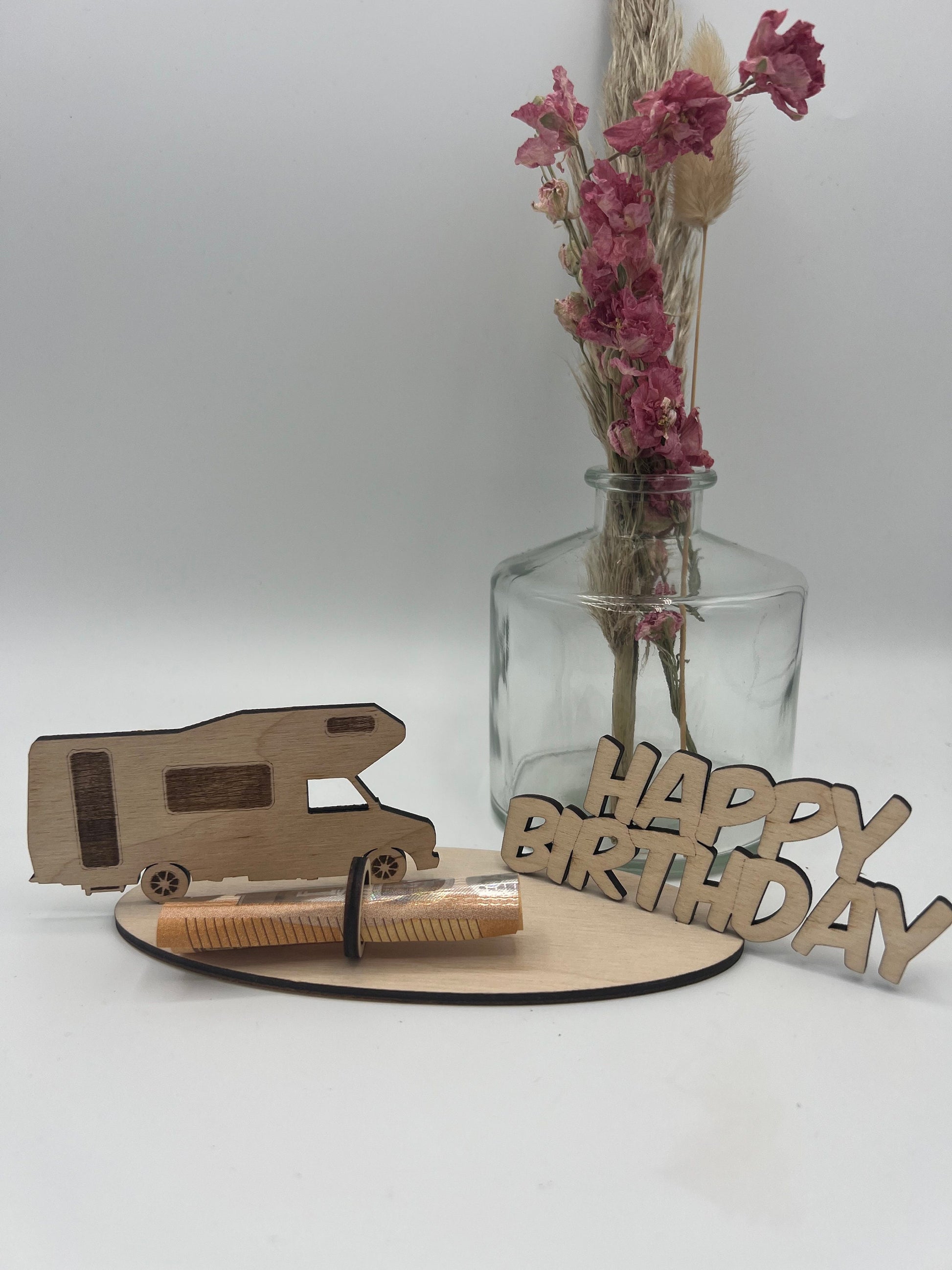 Geldgeschenk aus Holz in Form eines Campers mit der Aufschrift "Happy Birthday", dekoriert mit Geldschein auf einer Holzplatte und Blumen im Hintergrund.