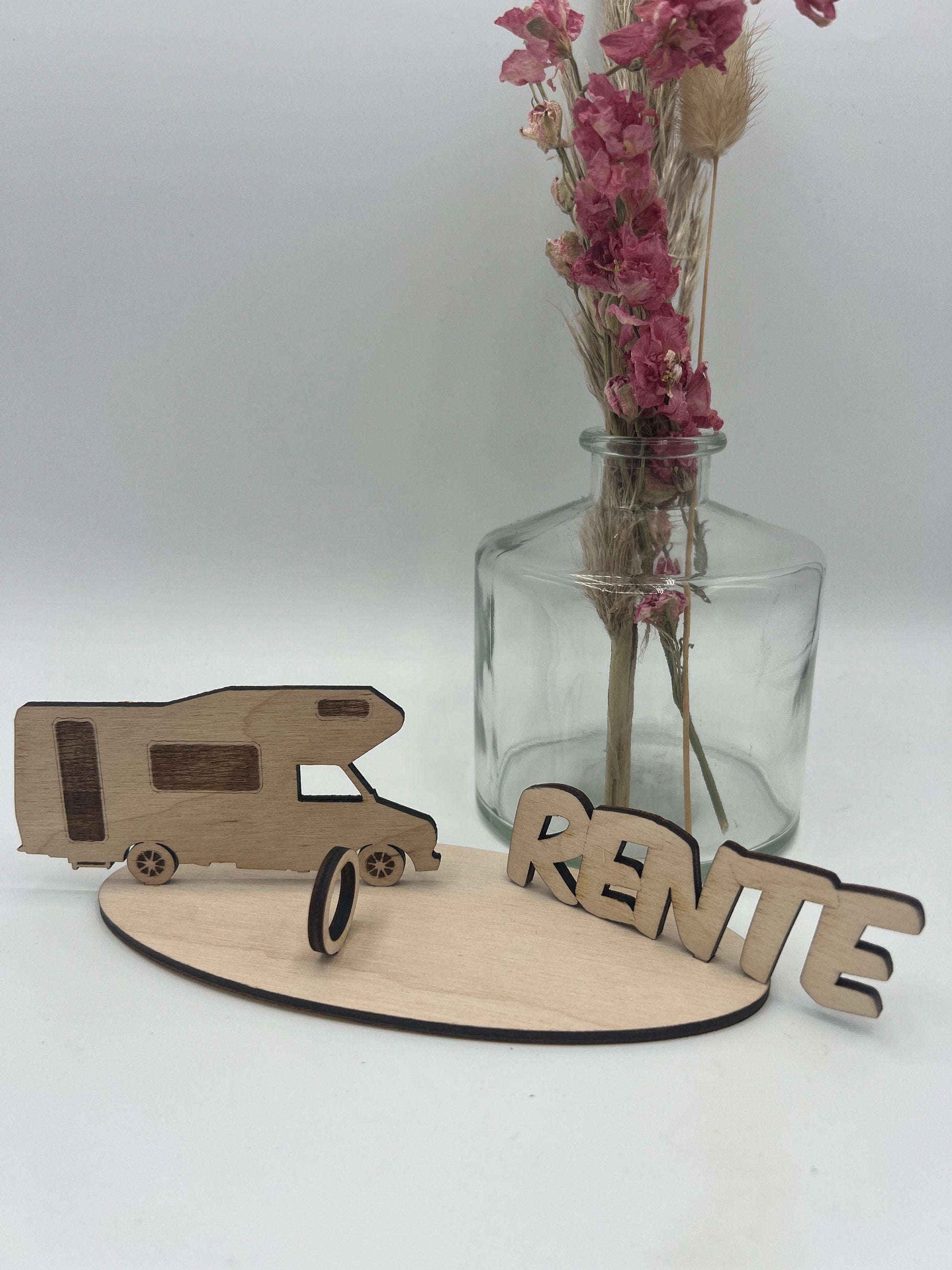 Geldgeschenk aus Holz in Form eines Campers mit der Aufschrift "Rente", dekoriert mit Geldschein auf einer Holzplatte und Blumen im Hintergrund.